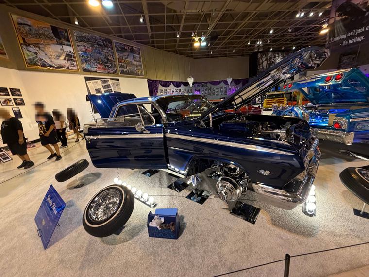 Dark blue lowrider with wheels off and the hood open on a show floor